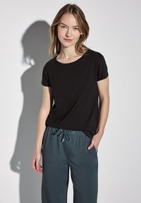 Černé tričko s krátkým rukávem z měkkého materiálu, doplněné volnými kalhotami na šňůrku v barvě teal. Má kulatý výstřih a jemné postranní rozparky.