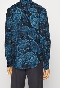 Langemouwshemden in marineblauwe stof met een groot blauw bloemenpatroon. Bevat knopmanchetten en een gebogen zoom. Textuur lijkt glad.
