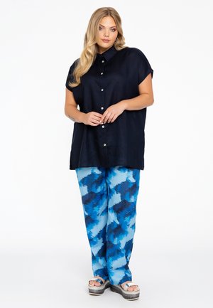 Marineblaues kurzärmliges Hemd mit Knöpfen, kombiniert mit blauen gemusterten Hosen, die Wellenlinien zeigen, getragen mit Plateausandalen.