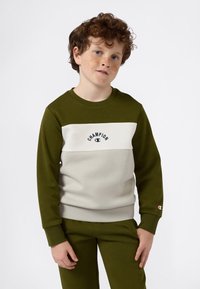 Niño con cabello rizado rojo y pecas, vistiendo un sudadero verde y beige de Champion y pantalones verdes a juego, de pie contra un fondo blanco.