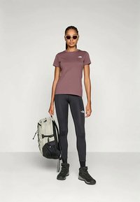 Naine, kes kannab prille, mauve värvi T-särki, musti retuuse ja musti matkasaapaid, hoiab käes heledat halli ja musta seljakotti.