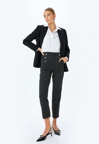 Schwarzes Blazer über einem weißen Hemd, kombiniert mit hochtaillierten Hose mit Schwarz-Weiß-Streifen und großen Knöpfen sowie spitzen schwarzen Absätzen.