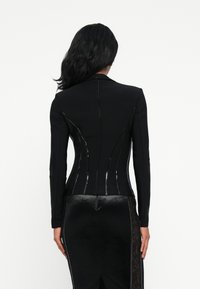 Γυναίκα με μακριά μαύρα μαλλιά που φοράει ένα εφαρμοστό μαύρο σακάκι και μαύρη φούστα με δαντελένια επένδυση, ιδωμένη από πίσω σε απλό λευκό φόντο.