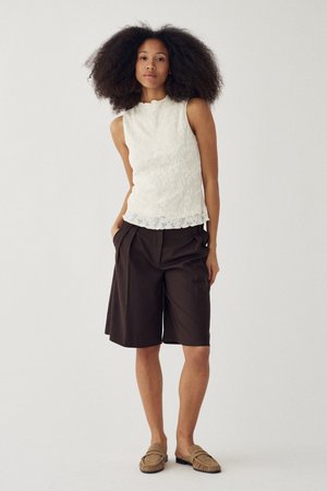 Nő dús, göndör hajjal, ujjatlan fehér felsőt, laza barna térd-length rövidnadrágot és világosbarna bebújós cipőt viselve, egyszerű háttér előtt áll.