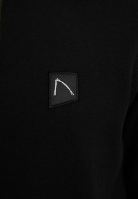 Černý svetr s měkkou strukturou. Obsahuje černý čtvercový záplatu s bílým prošívaným designem. Detailní záběr na látku a logo.