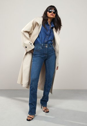 Žena v slnečných okuliaroch, ktorá má na sebe béžový trenčkot cez modrú džínsovú košeľu a rifle, stojaca v sebaistom postoji s jednou rukou v vrecku.