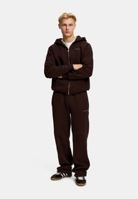 Brun zip-up hettegenser og matchende joggebukser laget av mykt stoff. Har logo-detaljer på brystet og buksene. Brukt sammen med svarte og hvite joggesko.
