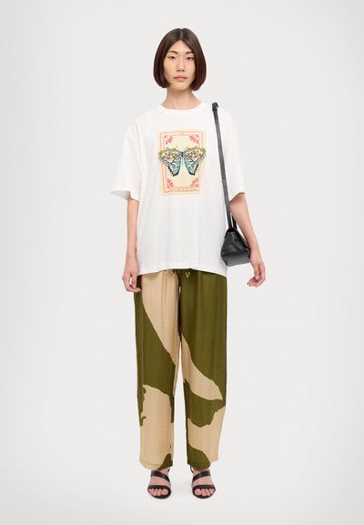Camiseta blanca de gran tamaño con un gráfico de mariposa, combinada con pantalones anchos de patrón en verde olivo y beige, y sandalias negras tipo slider.
