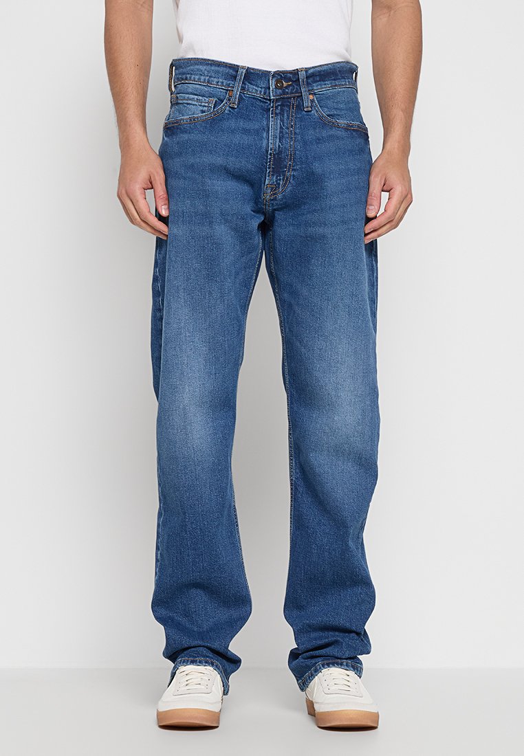 Blaue Jeans aus Denim mit geradem Schnitt, mittlerem Waschumfang, Fünf-Taschen-Design und kontrastierenden Nähten. Getragen mit einem weißen T-Shirt und hellen Sneakers.