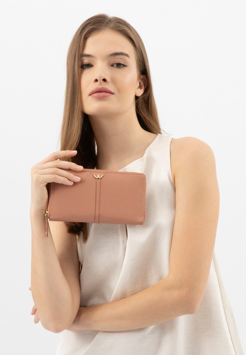 Giovane donna con capelli lisci castano chiaro tiene un portafoglio rosa con zip, indossando una canotta bianca sporca, su uno sfondo bianco semplice.