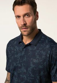 Námořnické modré polo tričko vyrobené z hladkého materiálu s jemnou texturou, dvoubarevným zapínáním na knoflíky a logem na hrudi.