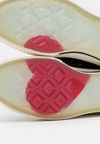 Gummisneaker mit durchsichtigen Sohlen, die ein Rautenmuster und einen roten herzförmigen Einsatz an der Unterseite aufweisen. Schwarzes Innenfutter.