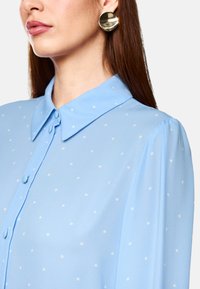 Light blue blouse with a collared neckline, button front, and white polka dots. Long sleeves with slight gathering at shoulders.