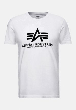 Hvit bomull t-skjorte med en svart trykt logo med et "A" og tre horisontale striper. Teksten inkluderer "ALPHA INDUSTRIES" og lokasjonsdetaljer.