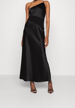 Mujer con vestido de satén negro sin mangas y de un solo hombro, ajustado en la cintura, y sandalias negras de tacón alto con tiras.