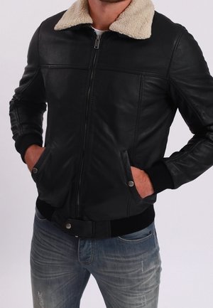 Man met een zwarte leren bomberjack met een beige schapenvachtkraag, handen in de zakken, gecombineerd met versleten blauwe spijkerbroek.