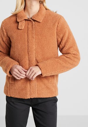 Femme portant une veste en polaire teddy marron avec un col et un pantalon noir, mains jointes à la taille.