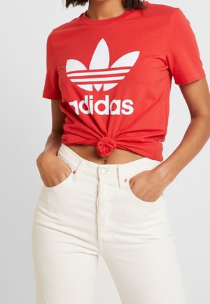 Kobieta w czerwonym krótkim T-shircie Adidas z logo, zawiązanym z przodu, w połączeniu z wysokim stanem kremowych dżinsów, ręce w kieszeniach.