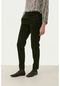 Olivgrüne Cordhosen im Slim-Fit mit seitlichen Taschen und einer leicht strukturierten Oberfläche. Kombiniert mit schwarzen Ballerinas und gepunkteten Strumpfhosen.