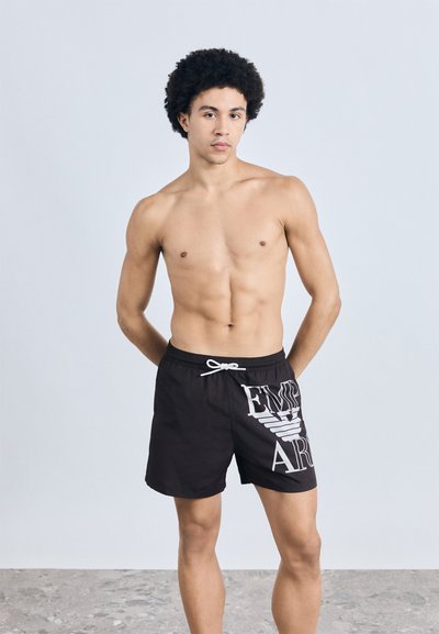 �Čierne plavkové šortky s bielym grafickým motívom na boku, s pásom so šnúrkou a ľahkým materiálom, zobrazené na modeli.