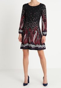 Vestido negro de manga larga, con patrones paisley rojos y blancos, acentos florales y una línea de cintura ajustada. Combinado con tacones altos azul marino.