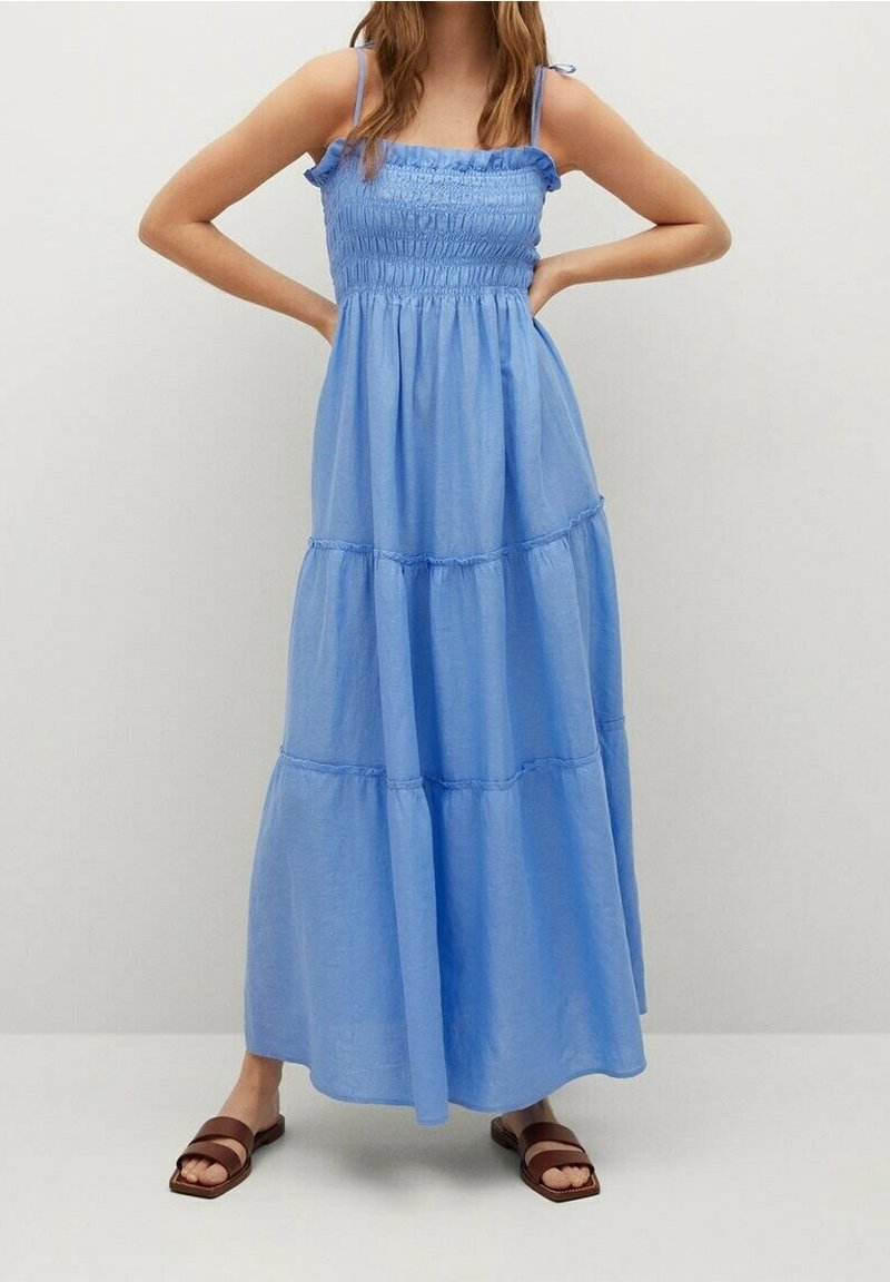 Femme portant une robe maxi bleue sans manches avec un corsage smocké et une jupe à volants, associée à des sandales plates marron.