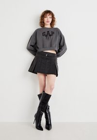 Grå croppad sweatshirt med "GAP"-logotyp och Mickey Mouse-tryck, matchad med en svart plisserad mini kjol och blanka svarta knähöga stövlar.