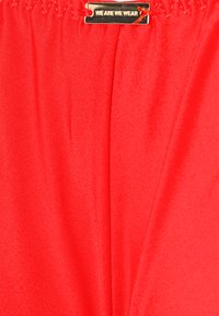 Roupa vermelha com tecido liso e brilhante. Apresenta uma etiqueta dourada com o texto "WE ARE WE WEAR" costurado na borda superior.