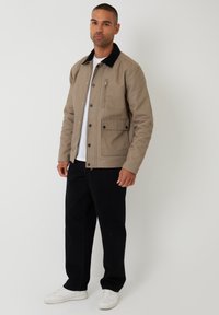 Beige jasje met zwarte kraag, ritssluiting aan de voorkant en drukknoopjes. Beschikt over zijzakken en een borstzak. Gekleed met zwarte broek en witte sneakers.