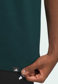 Dunkelgrünes Baumwoll-T-Shirt, kurze Ärmel, mit einem schwarzen Adidas-Tag am Saum. Glatte Textur, wird am unteren Rand gezogen.