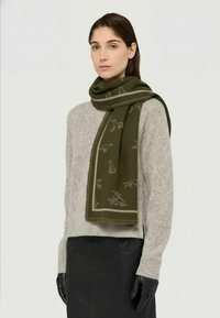 Sciarpa verde oliva con motivi animali e bordo a contrasto, indossata sopra un maglione lavorato a maglia grigio chiaro, abbinata a una gonna in pelle nera e guanti.