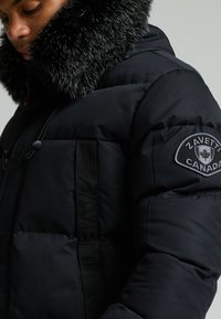 Puffjakke laget av svart stoff med en pluss svart pelsfôret hette, med et logopatch på ermene og en teksturert frontpanel.