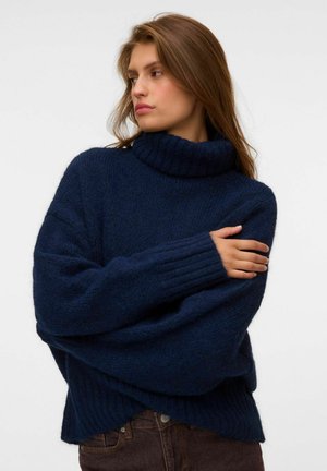 Jeune femme aux longs cheveux bruns porte un pull à col roulé épais bleu marine, regardant vers sa gauche, les bras croisés sur la poitrine.