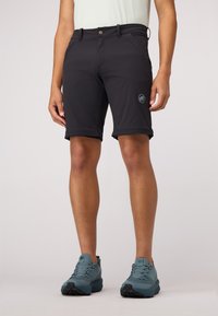 Shorts noirs en tissu extensible avec une fermeture à bouton à l'avant, des poches latérales et un logo sur la jambe gauche. Portés avec des chaussures bleues.