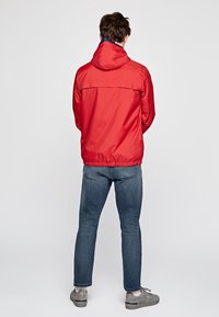 Rød vindjakke med hætte og elastisk kant, med en kontrastfyldt marineblå inderside. Bæres med blå jeans og grå sko.