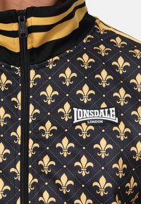 Melna zipa jaka ar dzeltenu fleur-de-lis rakstu, teksturētu audumu un izteiksmīgu baltu Lonsdale London logotipu uz krūtīm.