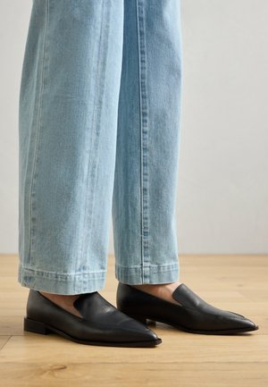 Chaussures en cuir noir à bout pointu et à enfilage, associées à un jean large bleu clair, sur un sol en bois.