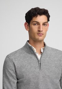 Grau zip-fester Pullover aus weichem Strickmaterial. Verfügt über einen Stehkragen und wird über einem weißen Hemd mit Kragen getragen. Einfaches und sauberes Design.