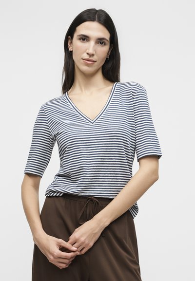 Γυναίκα με μακριά σκούρα μαλλιά, που φοράει μπλε σκούρο και λευκό ριγέ μπλουζάκι με V λαιμόκοψη και καφέ παντελόνι με κορδόνι, ενώ έχει τα χέρια της σφιχτά μπροστά.