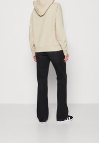 Felpa beige con cappuccio, vestibilità larga, abbinata a jeans neri a campana e sneakers nere con strisce bianche, con un focus sul comfort e l'abbigliamento casual.