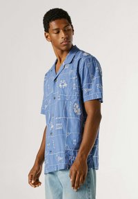 Jeune homme portant une chemise bleu à manches courtes avec des motifs blancs cousus et un jean bleu clair, regardant vers le bas à sa gauche.