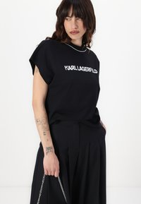 Melna kokvilnas T-kreklā ar īsām, sarullētām piedurknēm un baltu "KARL LAGERFELD" podziņu. Saskaņota ar augsto jostasvietu, melniem plisētajiem biksēm.