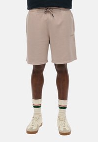 Shorts en coton beige clair avec taille élastique et cordon de serrage, dotés de poches latérales et d'un logo discret. Associés à des baskets crème et des chaussettes rayées.