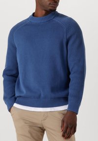 Maglione blu lavorato a maglia con un alto colletto a coste, maniche a coste e corpo liscio. Indossato sopra una camicia bianca e abbinato a pantaloni chiari.