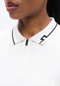Λευκό πόλο μπλουζάκι με μαύρο φερμουάρ και μαύρη λεπτομέρεια στο γιακά, με διακριτή υφή στον γιακά και μινιμαλιστικό στυλ.