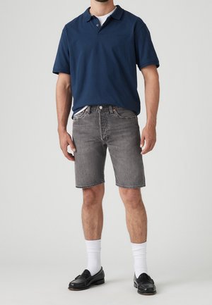 Marineblaues Polohemd, graue Jeansshorts, weiße Crew-Socken, schwarze Slipper. Lässiger Schnitt mit kurzen Ärmeln und kontrastierenden Nähdetails.