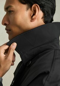 Guler de jachetă neagră cu detalii de cusătură triangulară, prezentată în momentul în care este ajustată de o mână. Textură de material neted, vedere detaliată de profil.