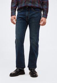 Άνδρας που φοράει σκούρα μπλε τζιν με ίσ ια γραμμή, μαύρα δερμάτινα μοκασίνια και πολύχρωμο καρό πουκάμισο με μακριά μανίκια, στέκεται μπροστά σε λευκό φόντο.