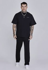 Crna oversized majica kratkih rukava s okruglim izrezom; kombinirana s crnim trenirkom s diskretnim logom; nošena s bijelim tenisicama.