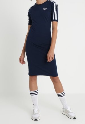 Femme portant une robe Adidas bleu marine avec des bandes blanches aux manches, des chaussettes blanches Adidas montantes et des baskets blanches, debout sur un fond uni.
